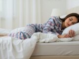 Junge Frau schlaeft entspannt in weissem Schlafzimmer – ideale Umgebung mit Nachttischlampe fuer gesunden Schlafrhythmus